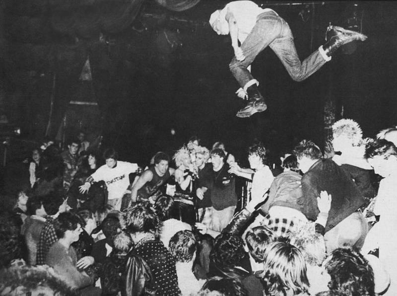A black-and-white photograph of a fan mid-air above a packed punk crowd at a club show.